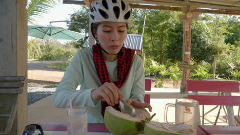 joven asiática con casco de ciclismo disfrutando de comer pulpa de coco tierna usando una cuchara en un pueblo en el norte de tailandia
