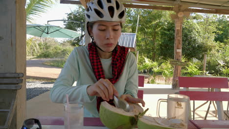 young asian woman wearing cycling helmet enjoying eating tender coconut pulp using a spoon in a village in northern thailand