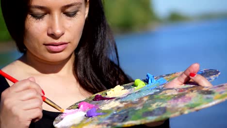 summer,outdoors, portrait of a beautiful forty years old brunette woman artist, close-up of a palette with paints, the woman artist mixes paints with a brush on the palette