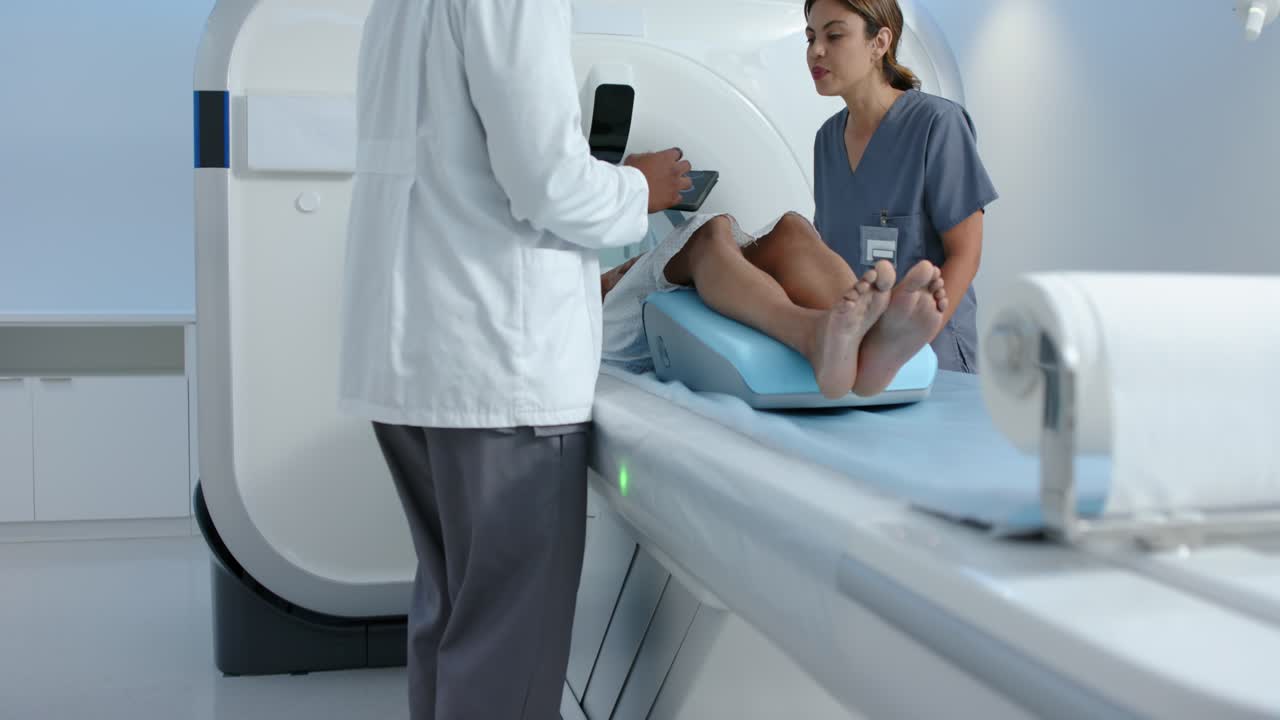 Uno staff afroamericano in camice e divisa da sala operatoria che utilizza un tablet controlla e modifica la posizione di un uomo sul cuneo per la TAC
