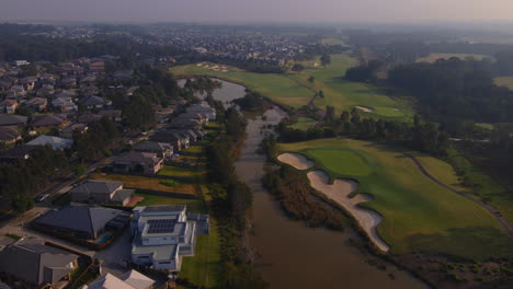 Aufsteigende-Luftaufnahme-Von-Großen-Australischen-Häusern-An-Einem-See-Und-Einem-Golfplatz-Belebter-Vorort-Mit-Verkehr