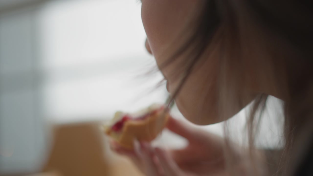 Tight over shoulder close up of young woman mid bite, holding pastry near lips while jaw, fingers, and loose hair move naturally in soft daylight inside calm interior, with woman, eating, pastry