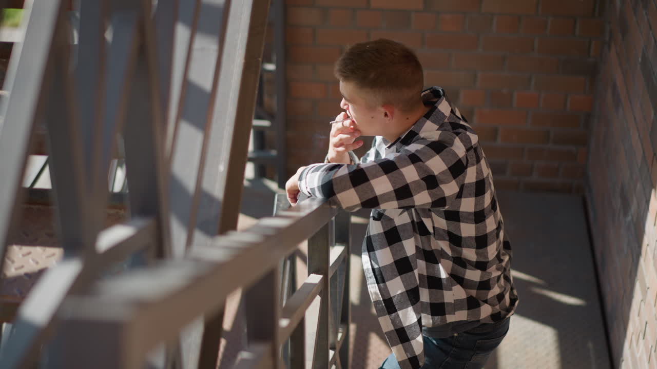 top down view of young student in checkered shirt and jeans leaning on iron rail smoking cigarette while sunlight filters through metal stair casting pattern shadows on weathered brick wall