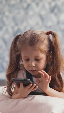 close up of cute little girl with blond long hair braided in pigtails in blue dress lies on floor in room under sunlight, child studies smartphone and video conference