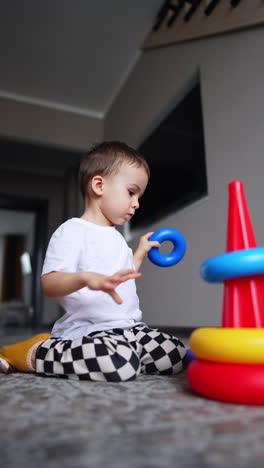 lovely baby boy sits on the floor disassembling the toy pyramid. cute toddler learning the shapes and sizes through the game. low angle view. vertical video.