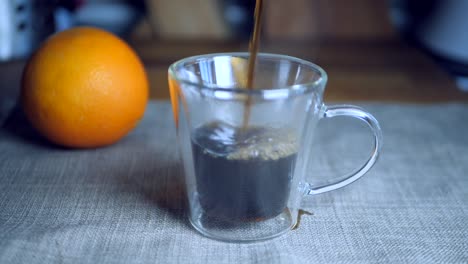 cámara lenta de café negro que se vierte en una taza de espresso italiana transparente sobre un trozo de tela gris junto a una naranja en el fondo