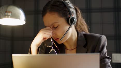 burnout and overworking concept - a woman takes off her glasses and massages the bridge of her nose while sitting in front of a laptop monitor.