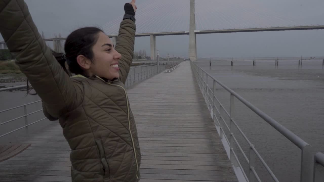 mujer joven sonriente corriendo en el muelle de madera con las manos levantadas