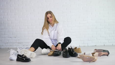 cheerful young woman sits on the floor choosing a pair of shoes. lady takes loafers from different pairs and compares them. white wall background.