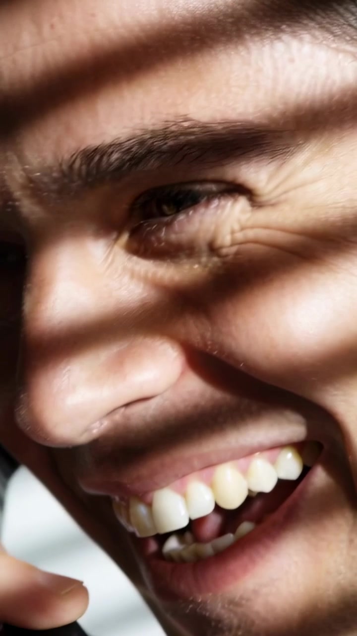 Vertical video: Bringing black handset to ear, laughing man speaking by blinds during call
