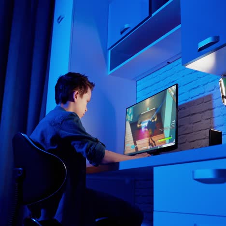 boy playing video game at home. concentrated gamer sitting at the desk in front of the computer and playing online games. blue light in the room.