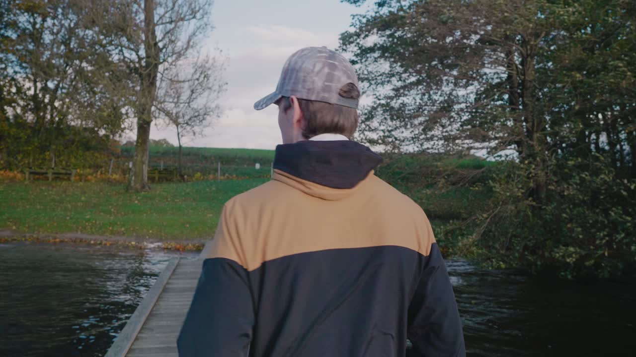 Young Tall Scandinavian Man With Cap Walks Down The Bridge on Gyllebo Lake on a Windy Autumn Afternoon - Tracking Medium Shot From Behind
