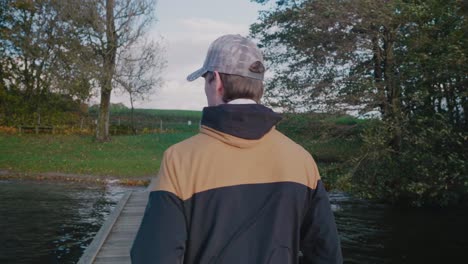 Young-Tall-Scandinavian-Man-With-Cap-Walks-Down-The-Bridge-on-Gyllebo-Lake-on-a-Windy-Autumn-Afternoon---Tracking-Medium-Shot-From-Behind