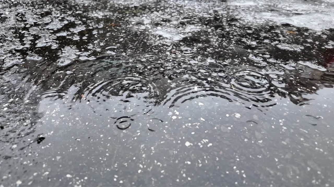 Raindrops hitting a dark asphalt surface, forming circular ripples and reflections during wet winter weather