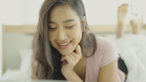 휴가 중 편안한 침실에서 침대에 누워있는 유쾌한 아시아 여성, 소셜 네트워크에서 친구들과 채팅하는 노트북을 사용하여 행복하게 미소 짓고, 원격 작업을하고, 온라인 쇼핑을하거나 소셜 미디어를 서핑합니다.