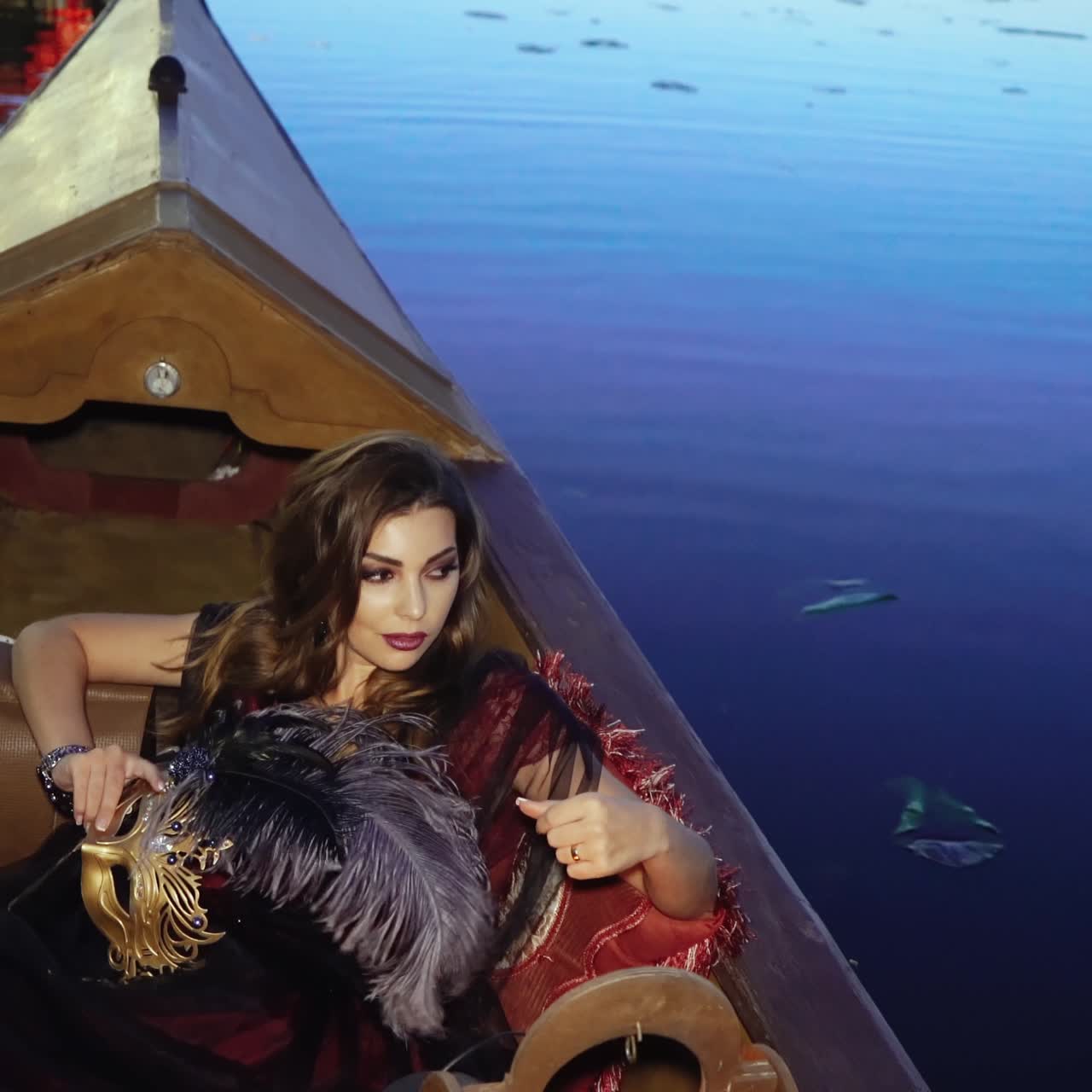 A pretty woman is lying in the gondola and stroking a purple feather from a mask in her hands on a blue background on the summer evening.