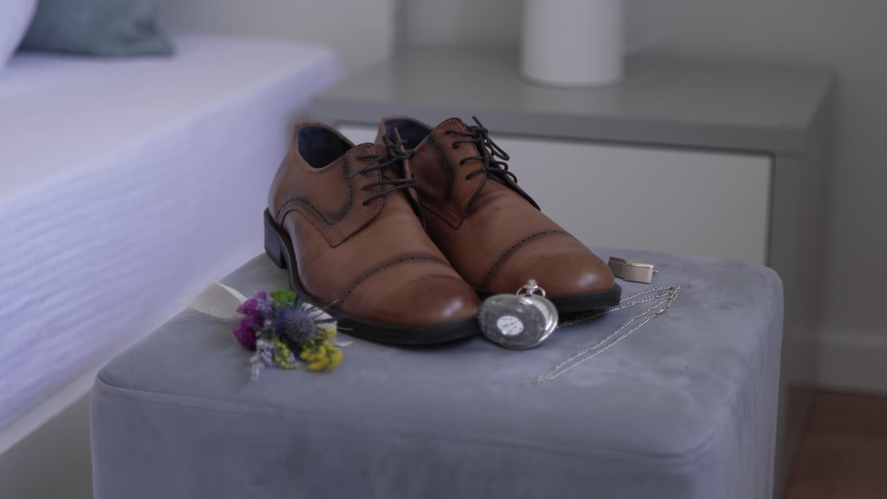Brown leather shoes, pocket watch, cufflinks, and boutonniere arranged on an ottoman
