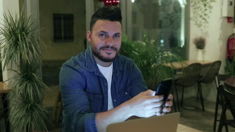 Handsome-man-using-smartphone-and-smiling-at-camera-in-cafe