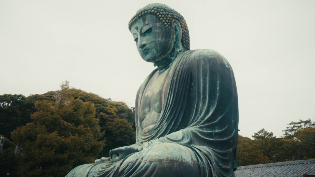 Una ripresa con tilt lento cattura l'imponente Grande Buddha di bronzo di Kamakura presso il Tempio Kotoku-in, Giappone, rivelando la sua patina invecchiata e la sua scala monumentale contro uno sfondo di collina boscosa.