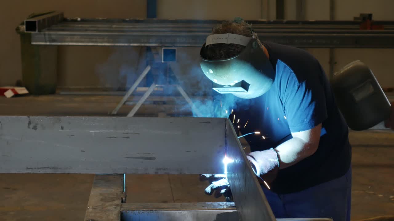 a man works welding metal in a metal products factory, the weld is blue light and sparks come out in all directions
