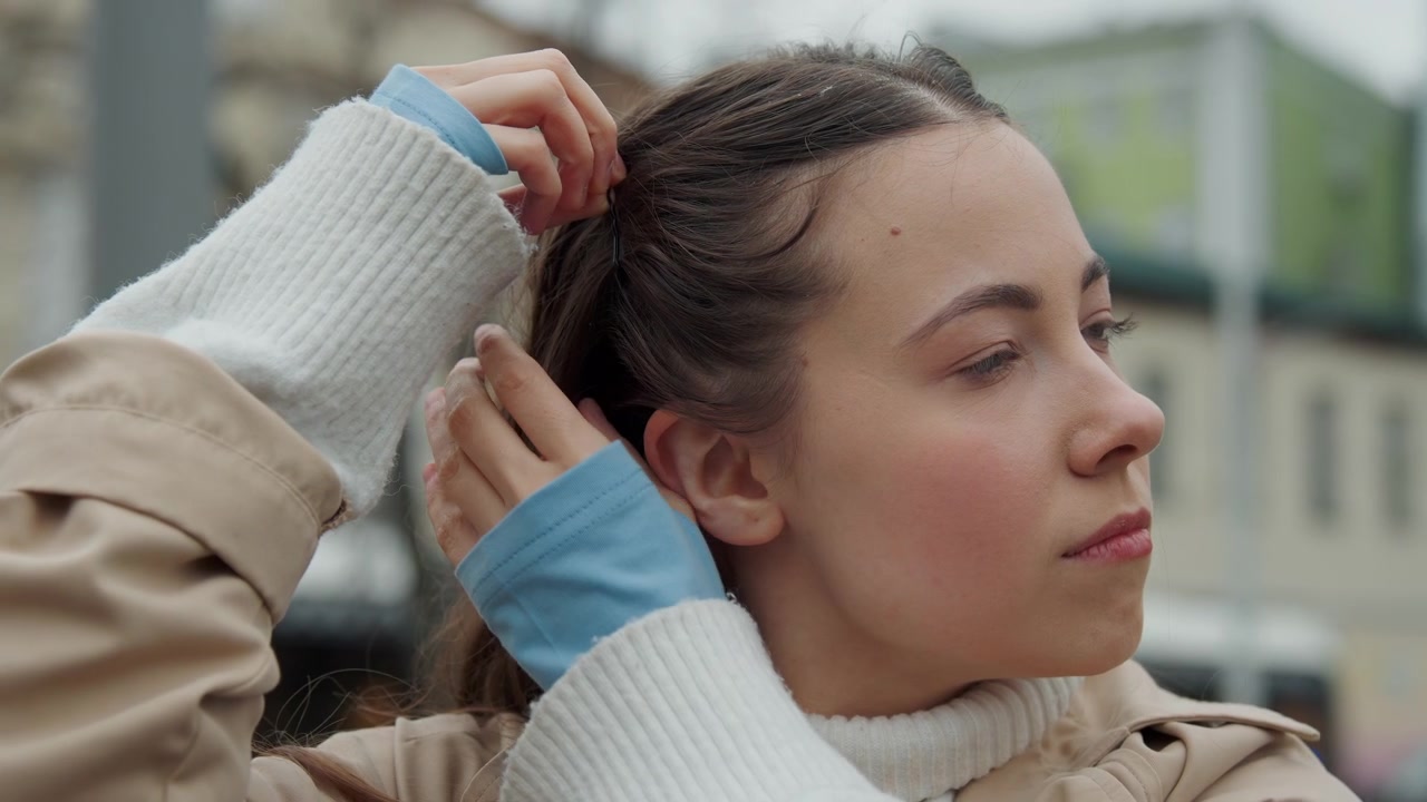 Primo piano di una giovane donna che si sistema i capelli in un ambiente urbano all'aperto. La donna indossa un cappotto beige sopra un maglione bianco con tessuto blu visibile ai polsini delle maniche. Lo sfondo mostra edifici sfocati