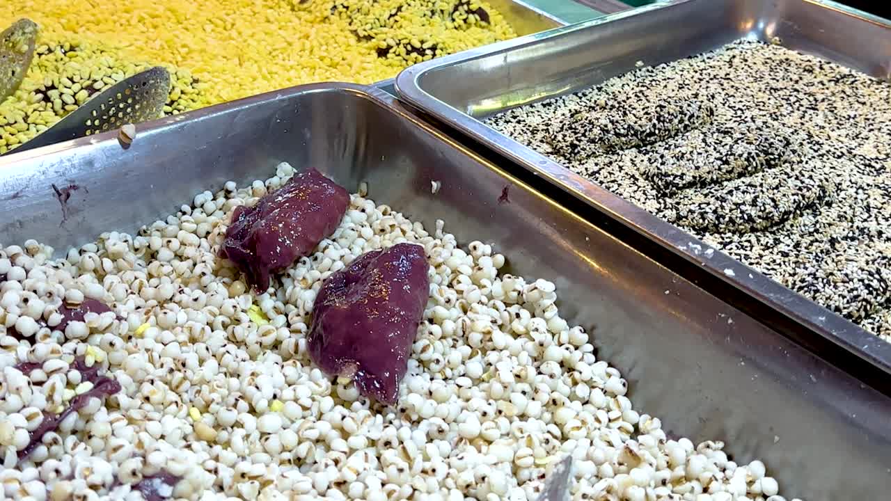 Close-up view of assorted grains and seeds in metal trays at a market stall.