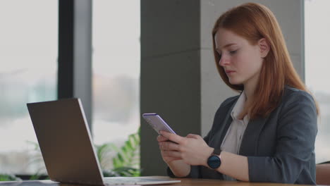 Hermosa-Joven-Mujer-De-Negocios-Usando-El-Teléfono-En-La-Oficina.-Mujer-Usando-Un-Teléfono-Inteligente-Y-Apoyada-En-Una-Ventana-Enviando-Mensajes-De-Texto-Enviando-Correos-Electrónicos-Planificando-Reuniones-Haciendo-Contactos-En-Línea-Navegando-Mensajes-En-Un-Teléfono-Móvil