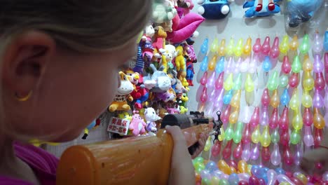 young caucasian girl shooting rubber bullet gun at rows of colorful balloons to win prizes at shooting range