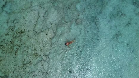 Young-Caucasian-woman-wearing-bikini-bathing-suit-floating-in-crystal-clear-green-ocean-water-looking-at-camera,-overhead-rising-aerial