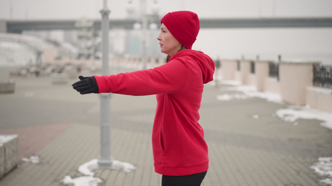 Woman in red hoodie and gloves performing hand stretches outdoors on snow-covered path with decorative metal fence, distant bridge, and moving car in misty atmosphere