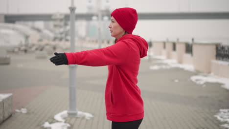 woman in red hoodie and gloves performing hand stretches outdoors on snow-covered path with decorative metal fence, distant bridge, and moving car in misty atmosphere