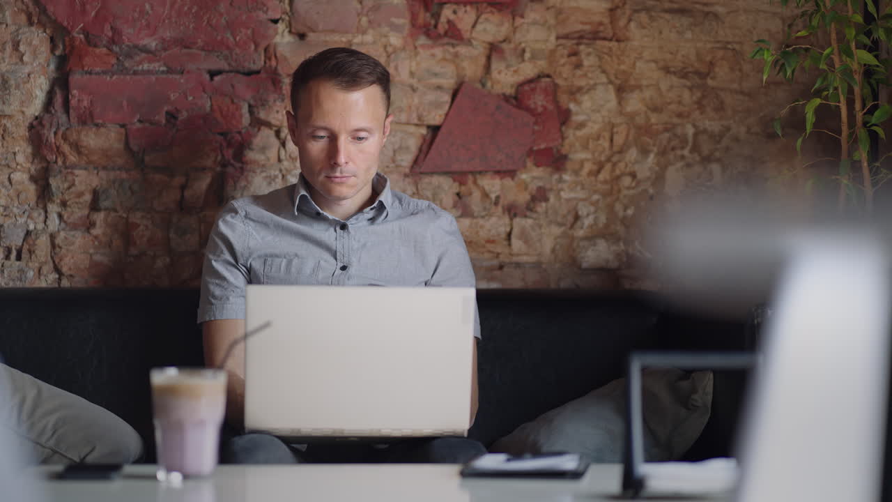 A young man in a shirt is sitting at a table with a laptop and typing on the keyboard. A student can study remotely. A businessman conducts his business remotely