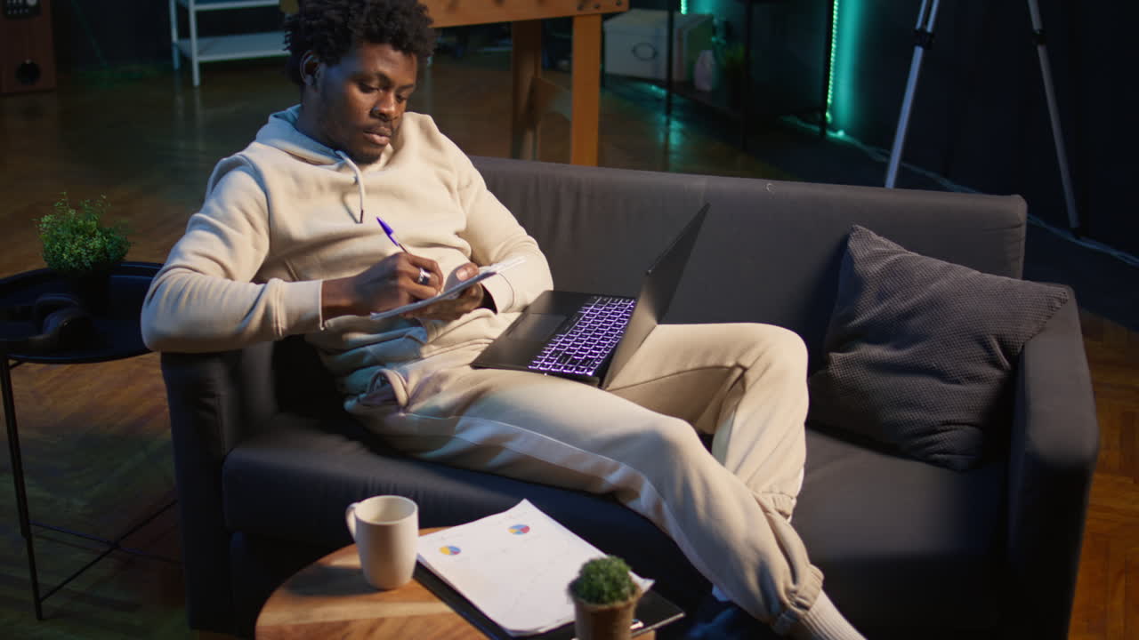 Casual person sitting On the sofa and reading research papers with analytics, multitasking with laptop at home. Man diving into professional or personal topics of improvement, independent. Camera B.