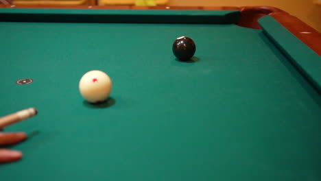 slow motion billiards person shooting solid black 8 ball in corner pocket on pool table with green felt stopping the cue ball in place with stun after practice strokes with cue stick close up