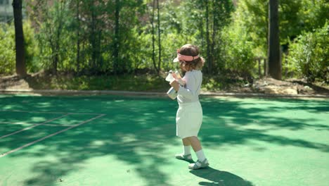 cute stylish 2-year-old child girl walking at badminton outdoor court on a sunny day wearing sport open top cap and holding water bottle - slow motion tracking shot