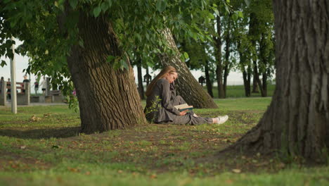 dama de ropa gris se sienta al aire libre bajo un árbol leyendo un libro con la mano en la página, ligeramente inclinada hacia adelante, pierna estirada, el fondo presenta vegetación y personas caminando en la distancia