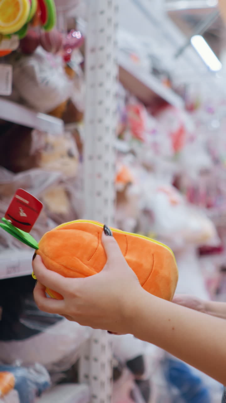 vista lateral de una mujer con una camiseta negra sosteniendo e inspeccionando un vibrante bolso de peluche naranja con detalles verdes en un centro comercial bien iluminado, examina cuidadosamente mientras navega por los estantes de juguetes y accesorios coloridos