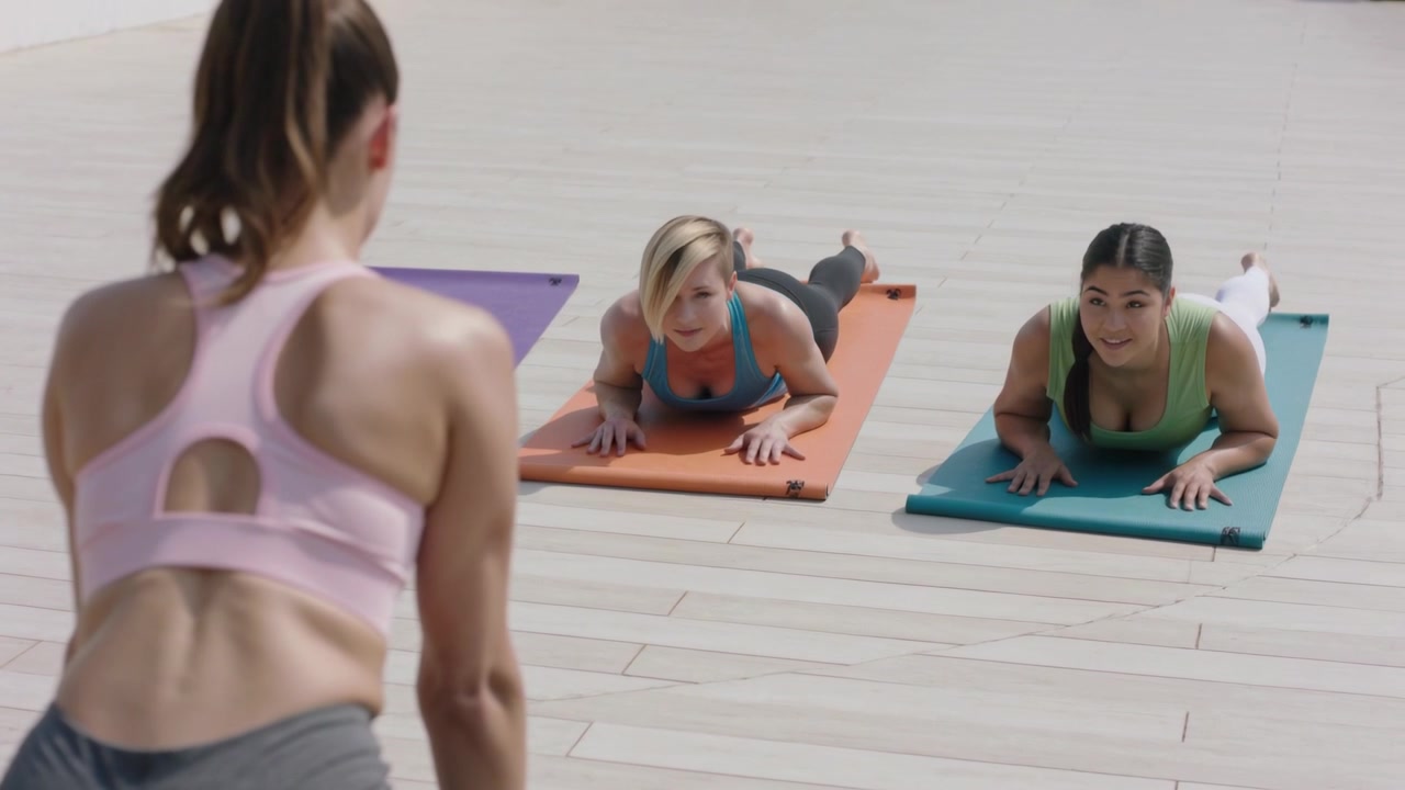Cueing instructor in pink sports bra, grey leggings guiding two pressing forearms on mats on deck