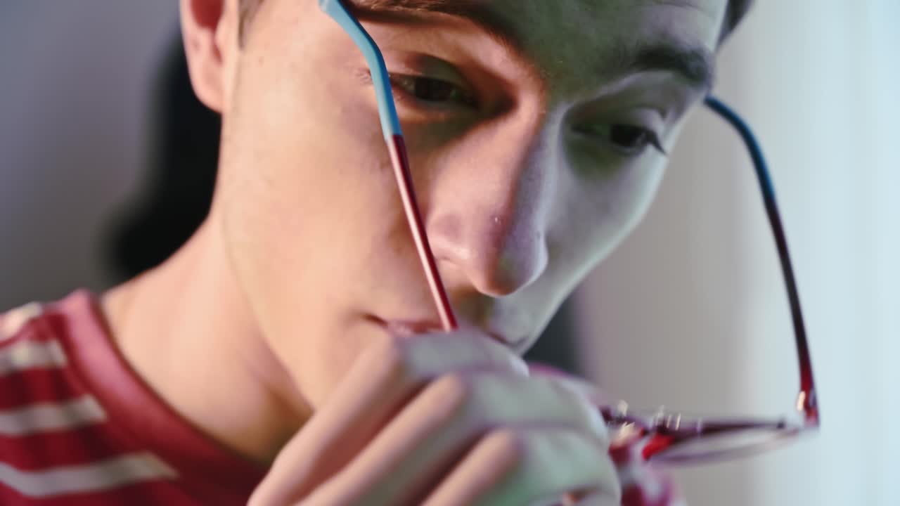 Tired young programmer in pink-striped sweater removes his red glasses on the eyes and massages his eyes. Green monitor reflection on his face. Close up
