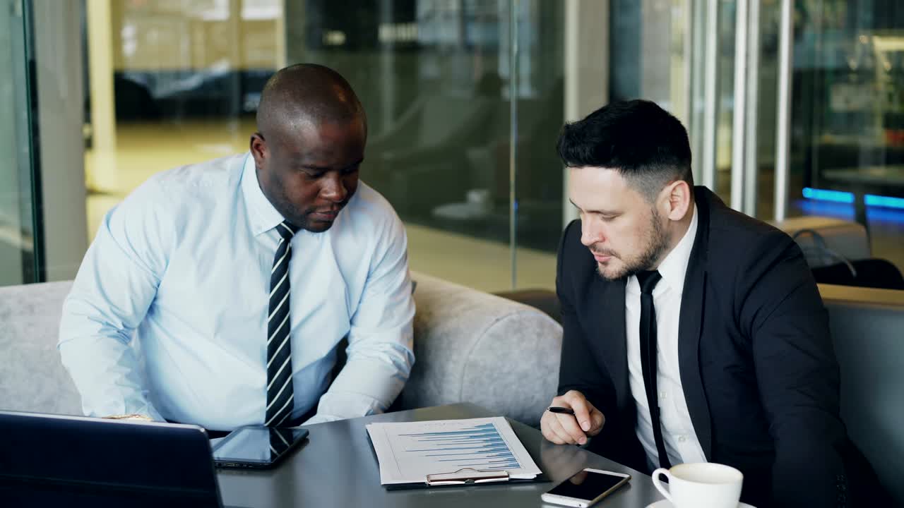 Two multi-ethnic partners in formal clothes sitting and discussing financial charts details while looking at notepad in glassy cafe during lunch time.