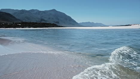 una desembocadura de la laguna que desemboca en el océano mientras las olas se deslizan suavemente y las montañas en la distancia en hermanus, sudáfrica.