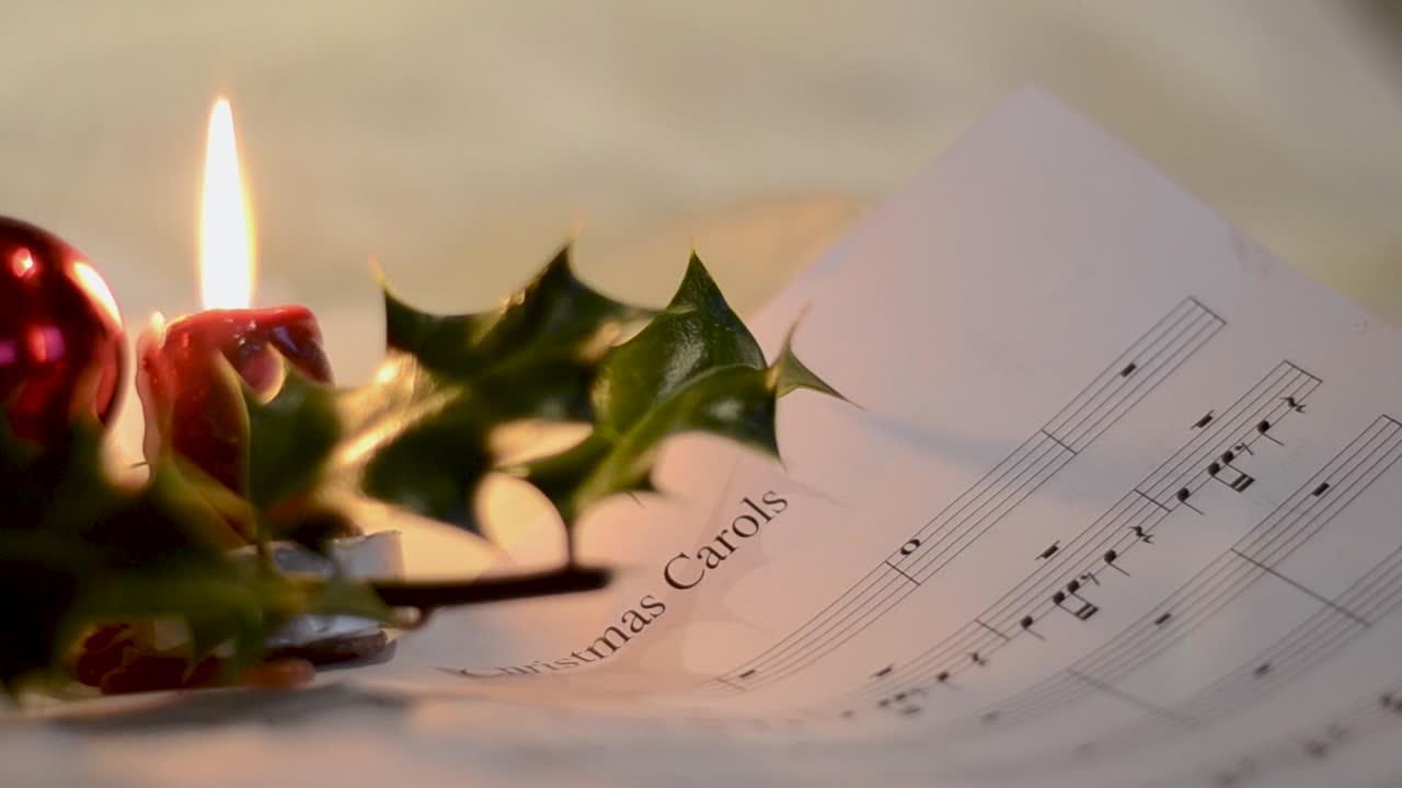 A close well focused shot of a white sheet contaning musical notations for christmas carol kept beside a chistmas holly plant and a burning small candle