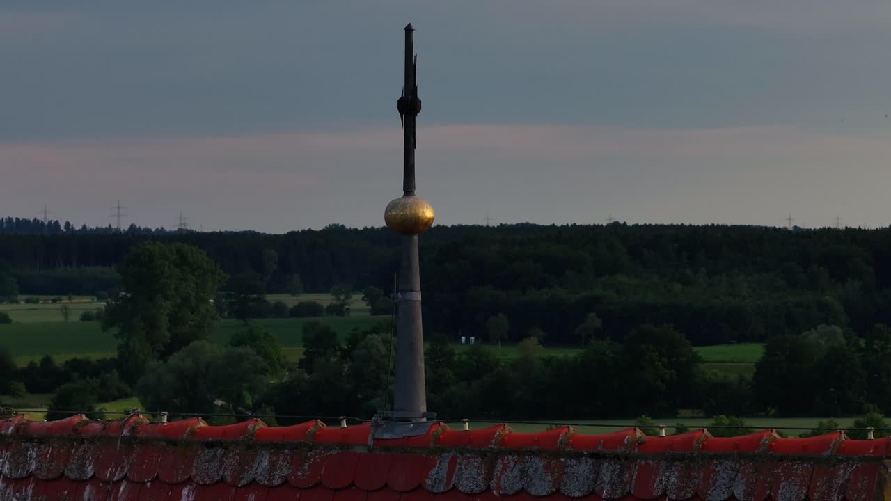 A weather vane on a rooftop overlooks a rural landscape with trucks on a nearby road at dusk