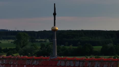 a weather vane on a rooftop overlooks a rural landscape with trucks on a nearby road at dusk