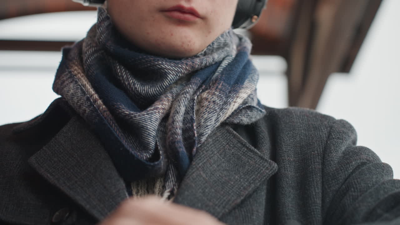 Jovem criador branco com fones de ouvido verificando o relógio, closes do rosto e das mãos, cachecol e casaco com neve, expressão pensativa, ajustando o relógio de pulso, em uma plataforma de inverno, pensativo.