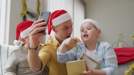 Kaukasischer-Vater-Und-Zwei-Söhne-öffnen-Geschenke,-Während-Sie-Zu-Weihnachten-Einen-Videoanruf-Auf-Dem-Smartphone-Führen