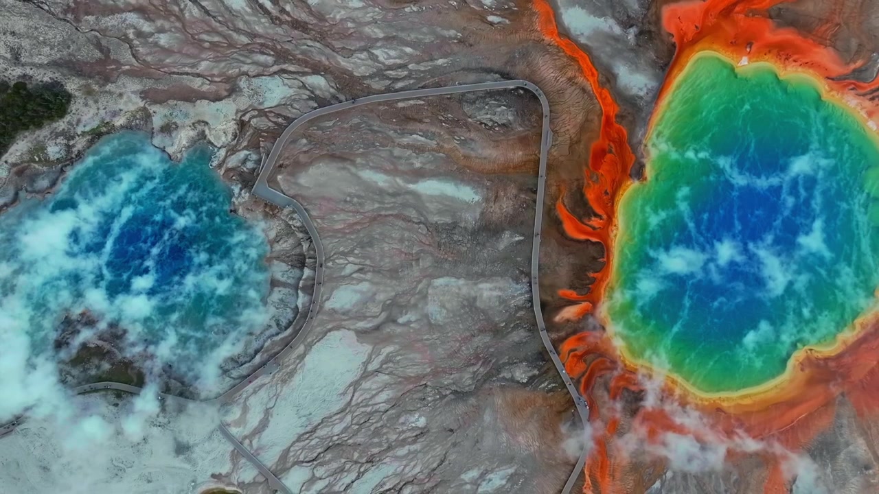 Flygfoto med drönare över Midway Geyser Basin och Grand Prismatic Spring i Yellowstone nationalpark, Wyoming, med gångbroar, två livfulla turkosa varma källor och vulkanisk termisk ånga som stiger uppåt