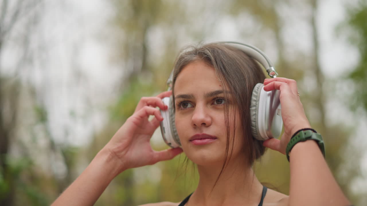 Close-up shot of beautiful young lady wearing headphones with focused expression while adjusting them on her head, she stands outdoors, blending into nature with a peaceful and concentrated demeanor