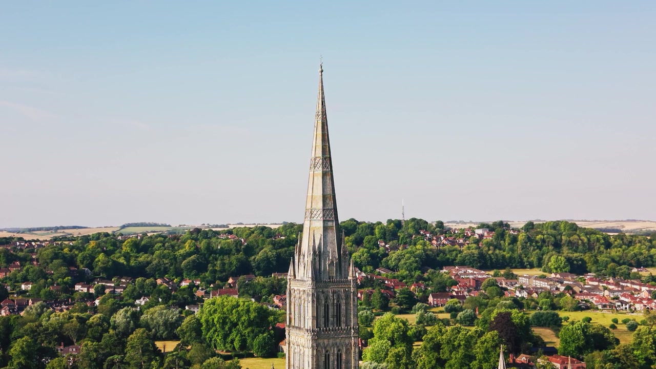 Vista drone della guglia della Cattedrale di Salisbury che si erge sopra alberi verdi, quartieri storici e la campagna circostante del Wiltshire in una limpida giornata estiva
