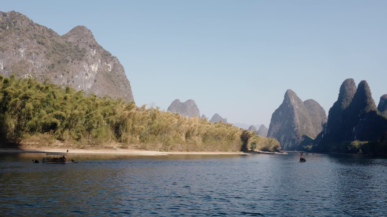 Small boats travel along the calm Li River as sharp karst mountains rise in the distance
